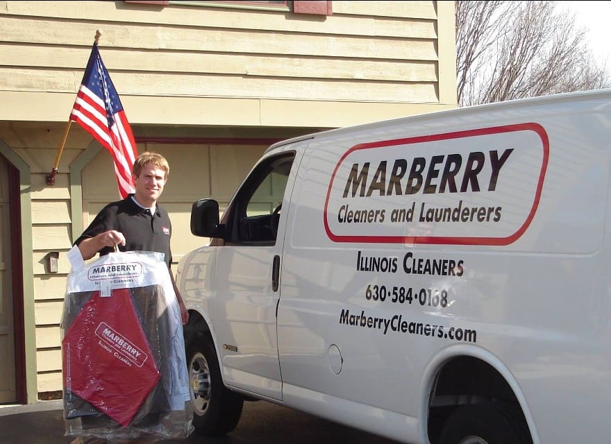 A man stands beside a Marberry Cleaners and Launderers van, holding cleaned clothes on hangers in front of a house with an American flag.