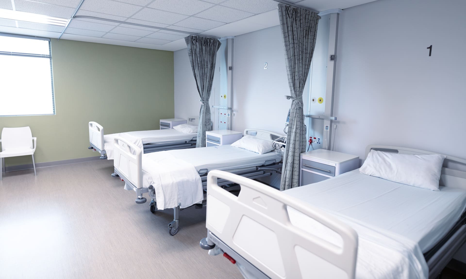 Three empty hospital beds with white linens are arranged in a row, separated by curtains, in a clean, well-lit hospital room.