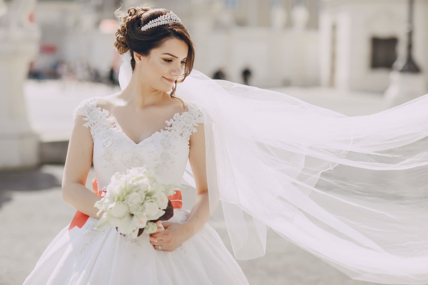 A bride in a white wedding dress and veil holds a bouquet of flowers, standing outdoors with soft sunlight in the background.