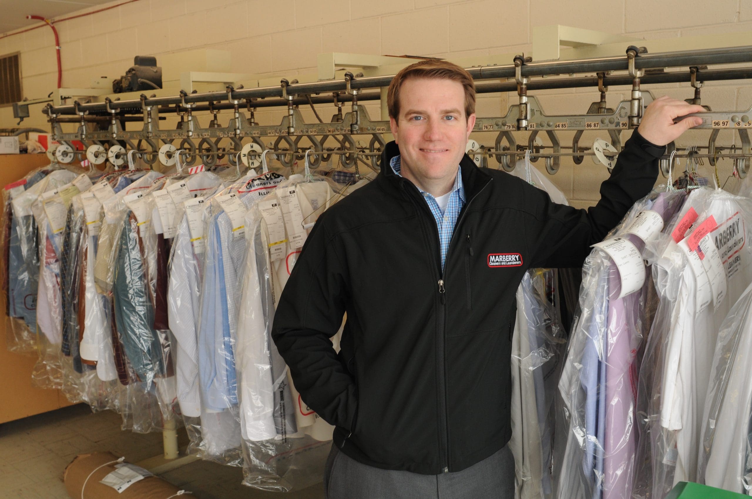 A man in a black jacket stands in a dry cleaning shop, surrounded by clothes hanging on a conveyor system, some covered in plastic.