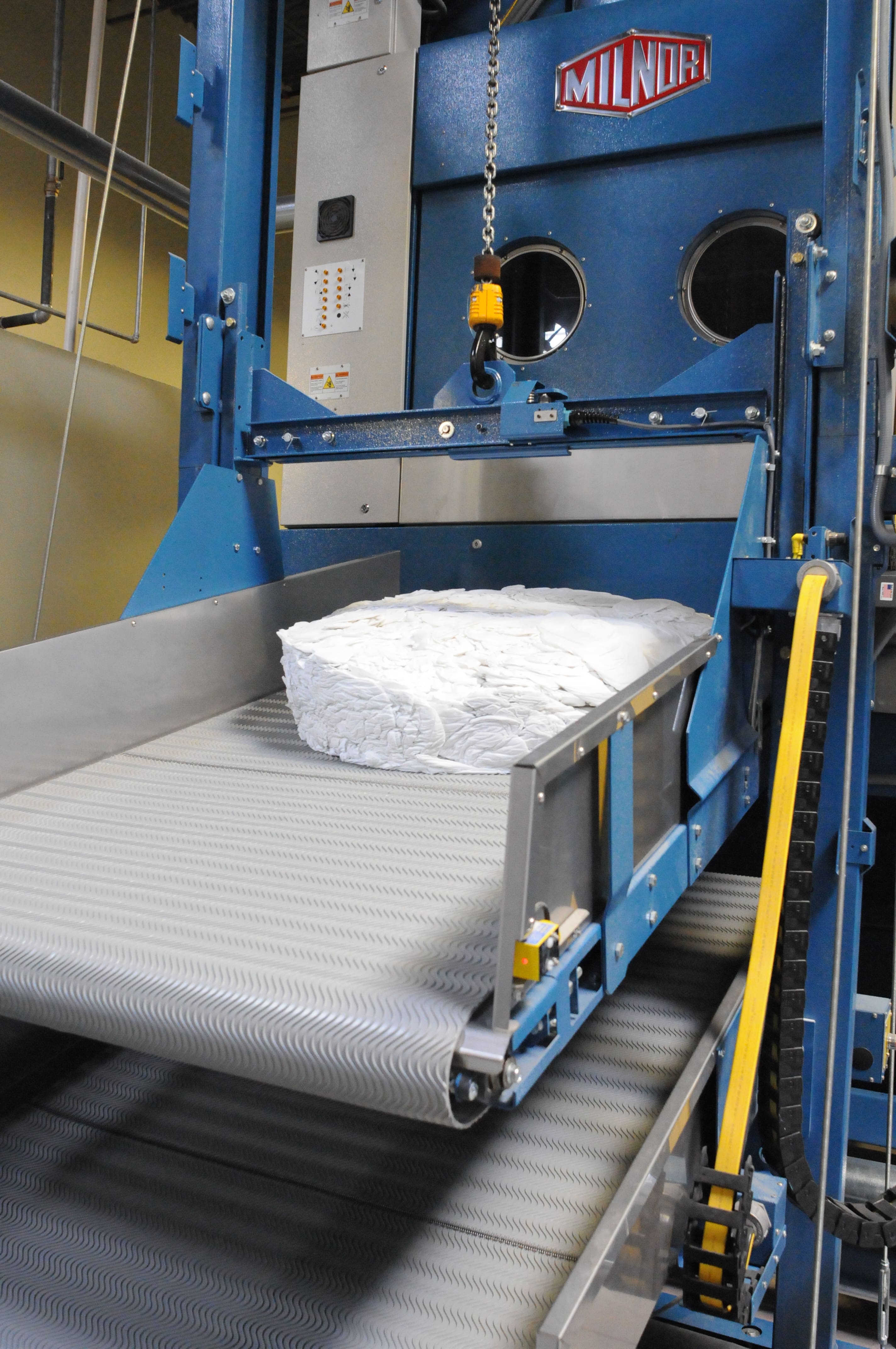 A large industrial washing machine unloads a stack of white linens onto a conveyor belt.