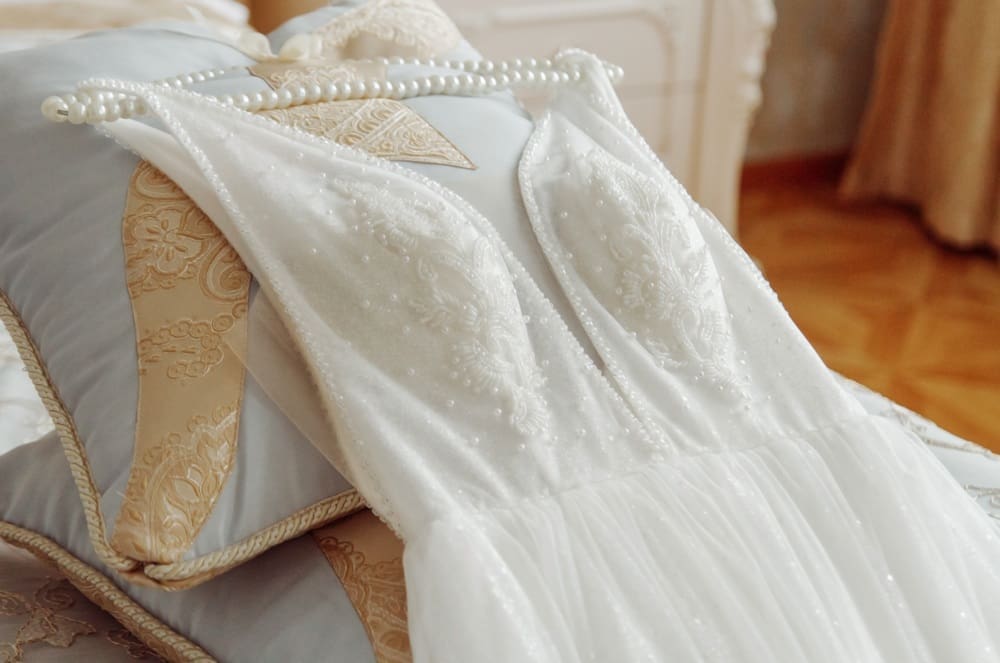 A white wedding dress with beaded details and a pearl necklace is laid out on a decorative pillow.
