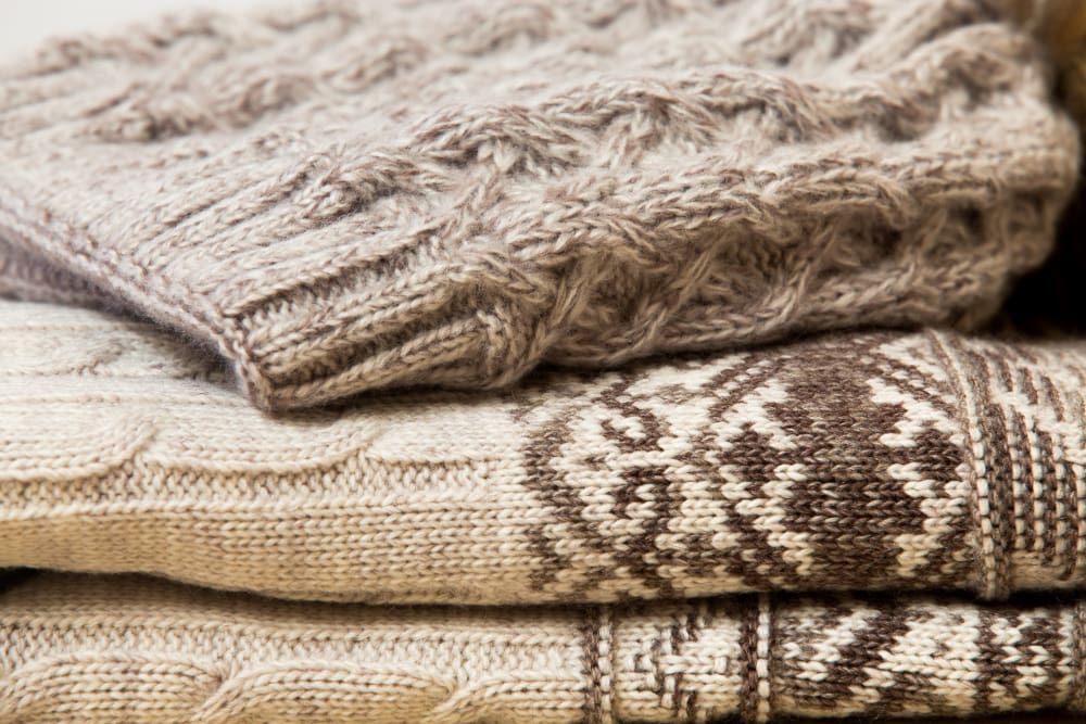 A close-up of three folded knitted sweaters in neutral colors, with visible patterns and textures, stacked on top of each other.