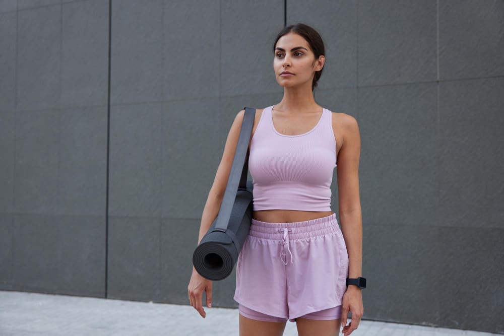 Woman in a pink sports outfit stands outdoors with a yoga mat over her shoulder, looking ahead, against a gray wall background.