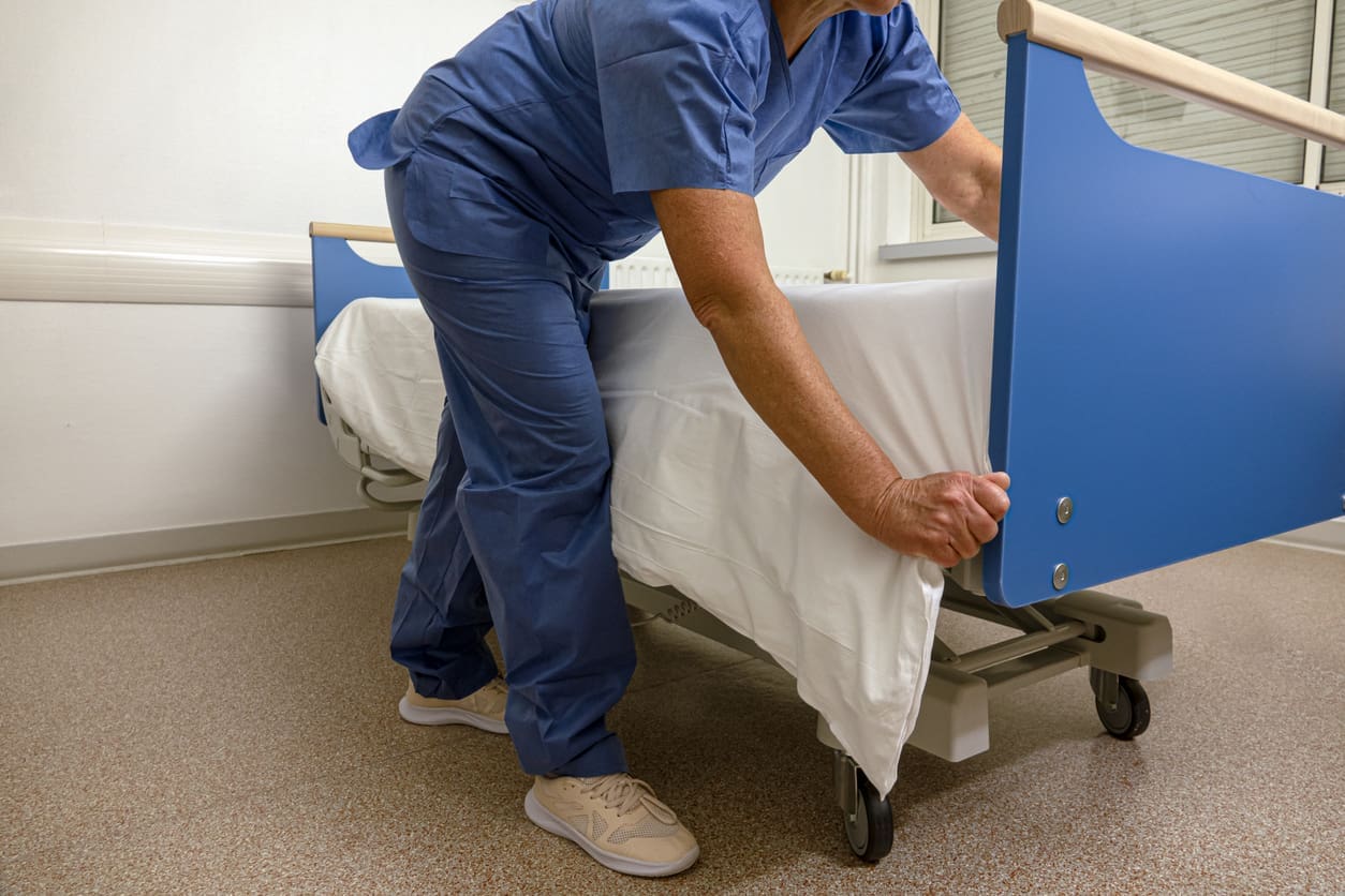 A healthcare worker in blue scrubs tucks a white sheet under a hospital bed mattress.