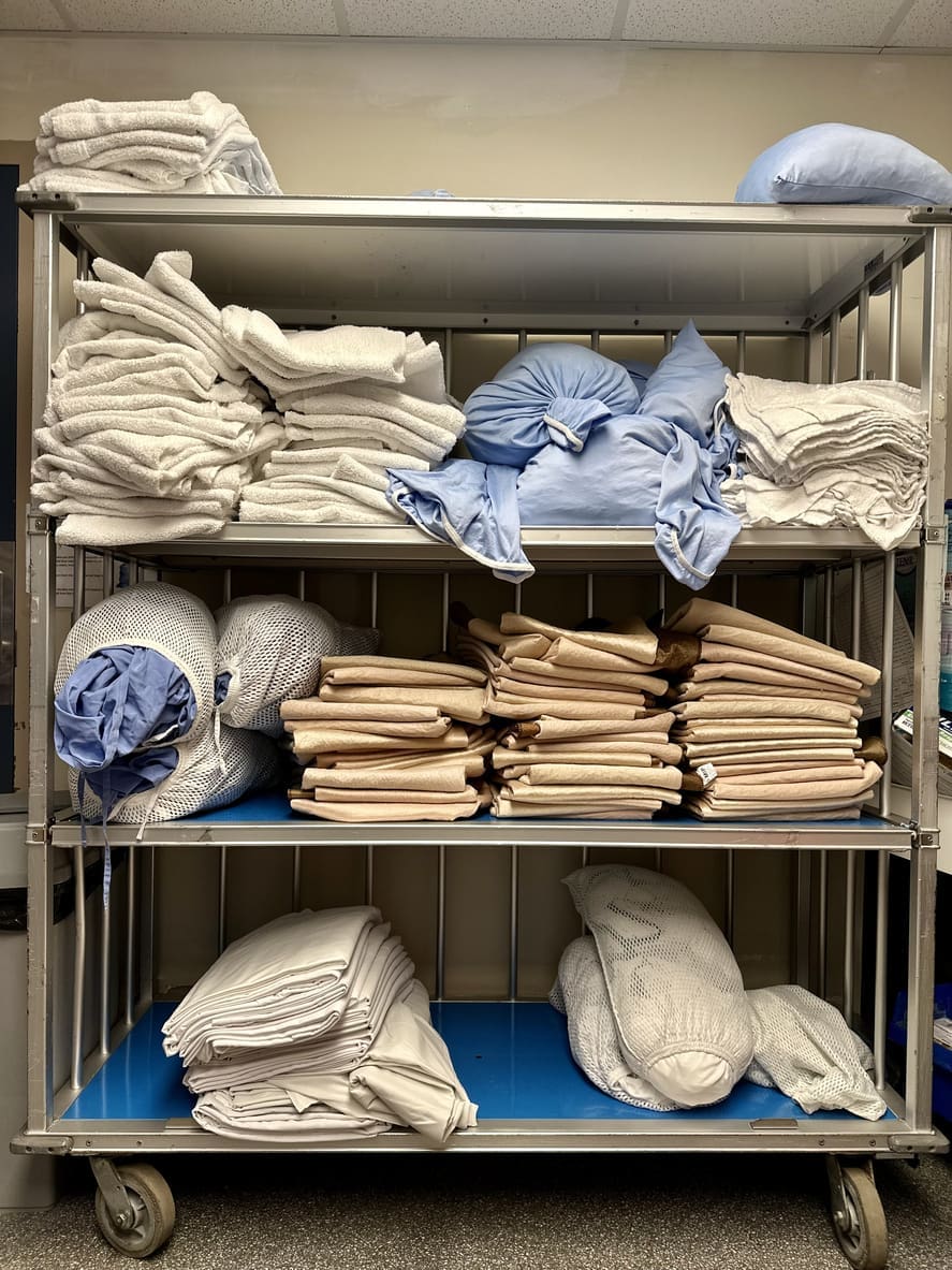 A metal cart with three shelves holding stacks of folded white and beige towels and blankets.